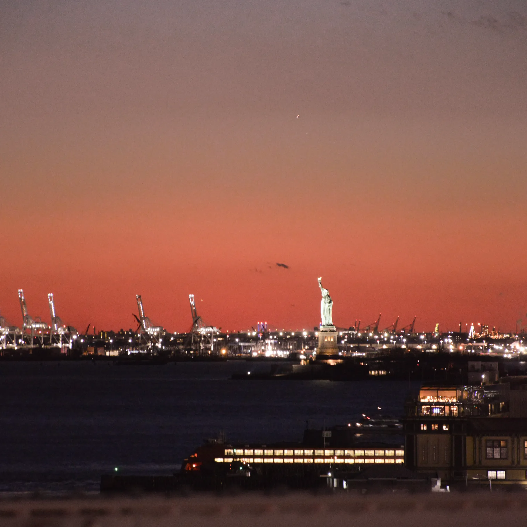 The Statue of Liberty against the last red of the sky, the port cranes of New Jersey behind.